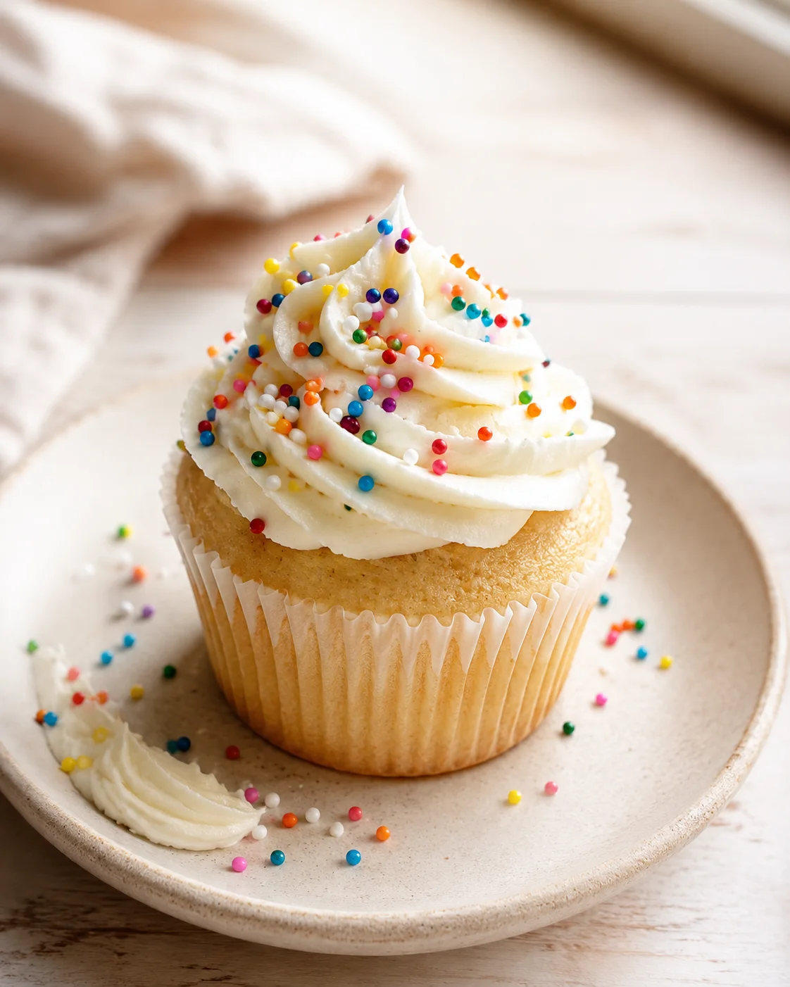 A vanilla cupcake with white frosting piped in a tall swirl, topped with rainbow sprinkles on a beige plate.