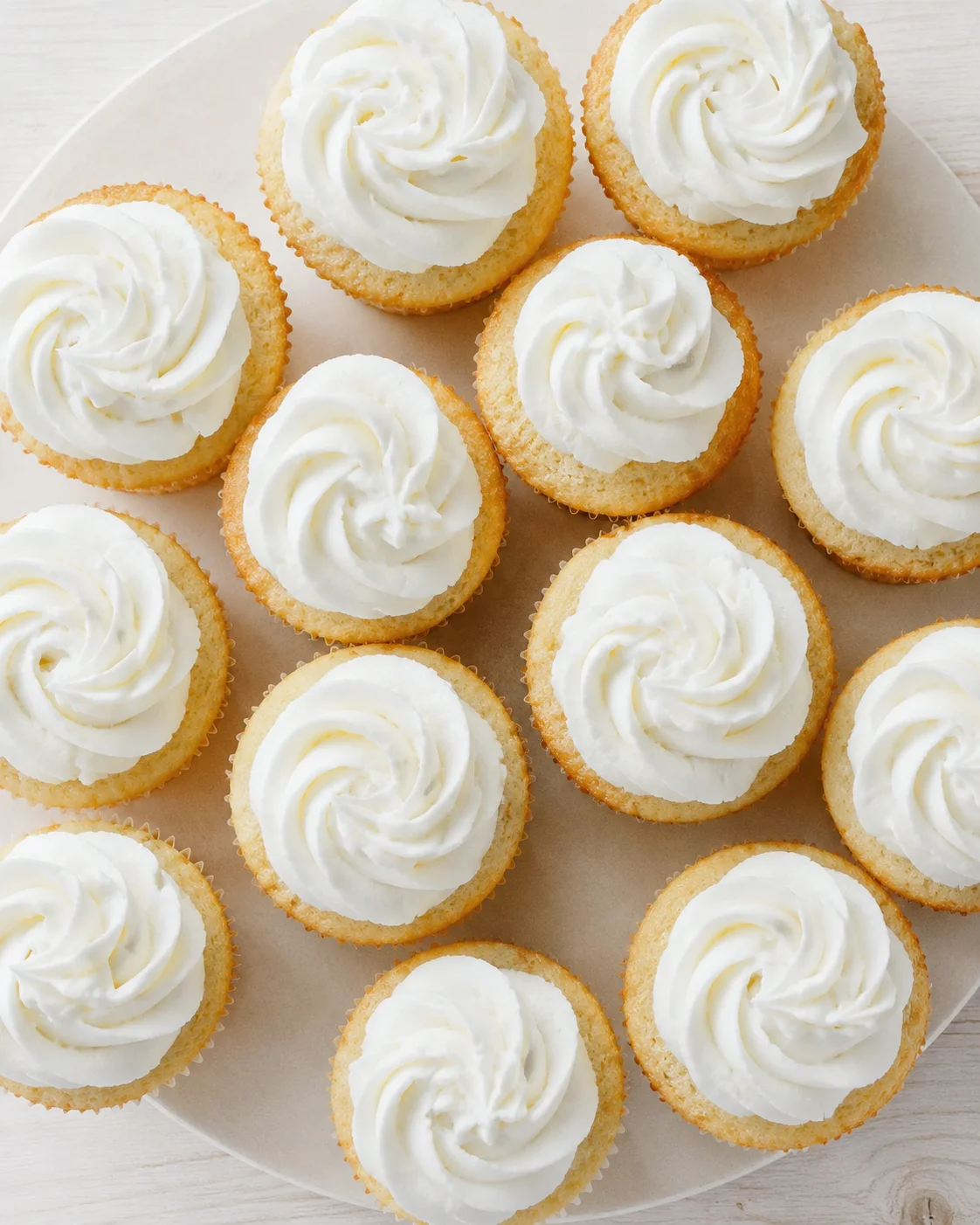 Plate of vanilla cupcakes with white buttercream frosting swirls on a light wooden background