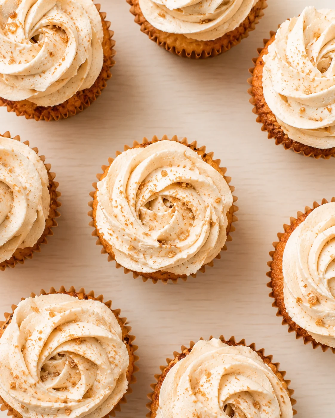 Top-down view of vanilla cupcakes with swirled cream-cheese frosting on a light wood background