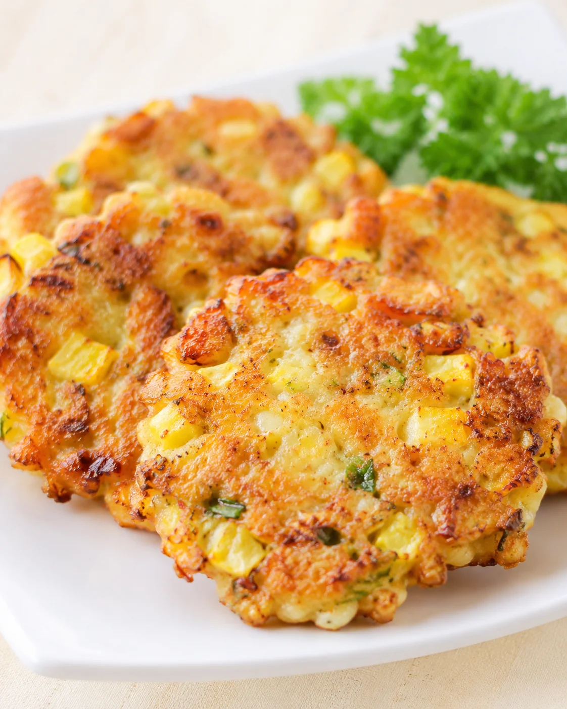 Golden-brown corn and potato fritters on a white plate with parsley garnish