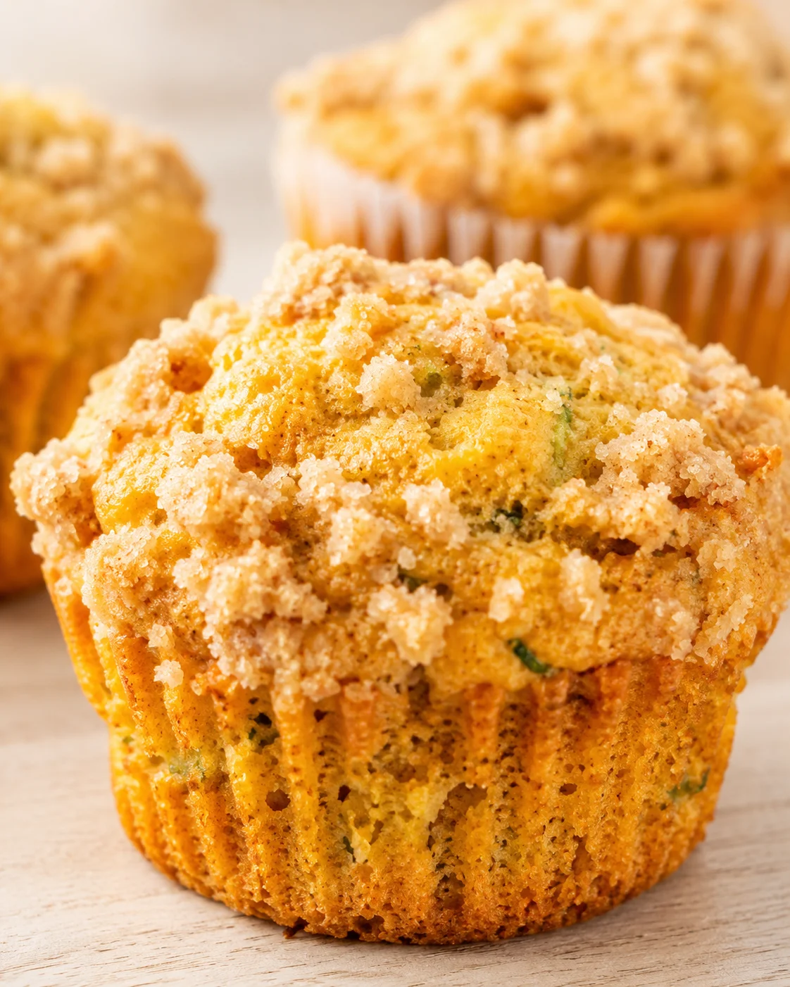 Golden crumb-topped muffins in paper liners on a light wooden surface.