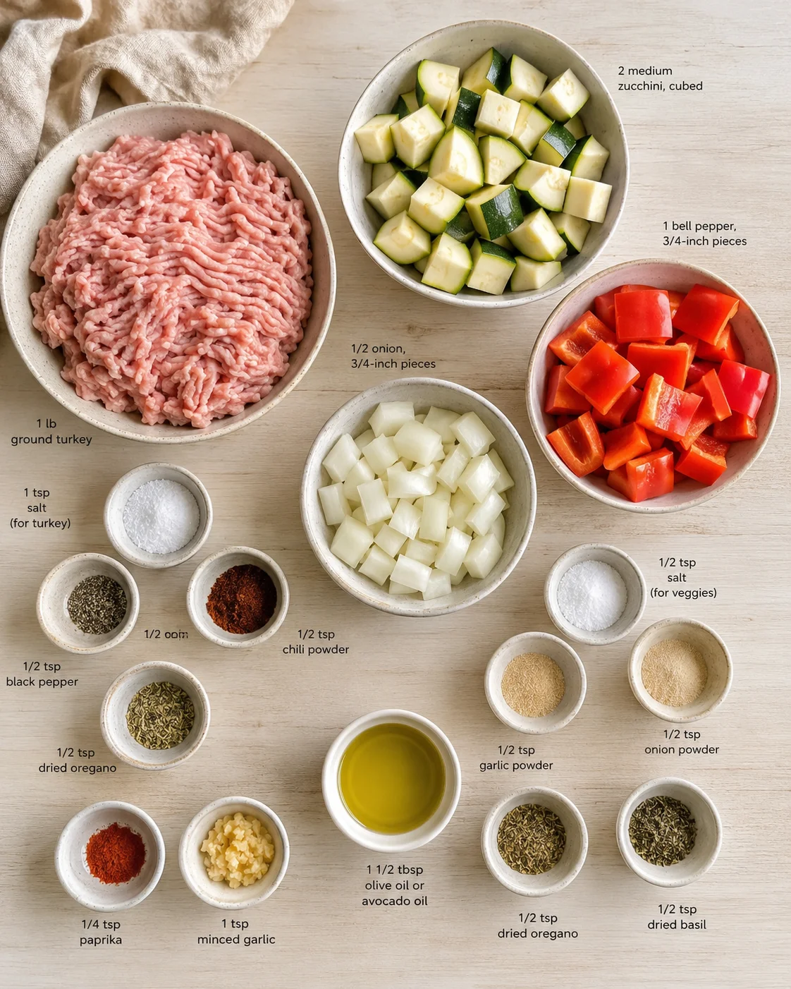 Overhead view of bowls with ground turkey, zucchini, onion, peppers, and spices on a light wooden surface.