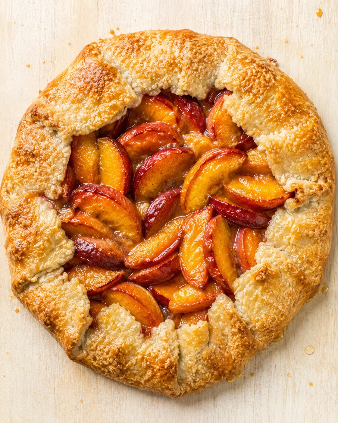A rustic peach-plum galette with a flaky golden crust and glossy fruit filling on a light wooden board.