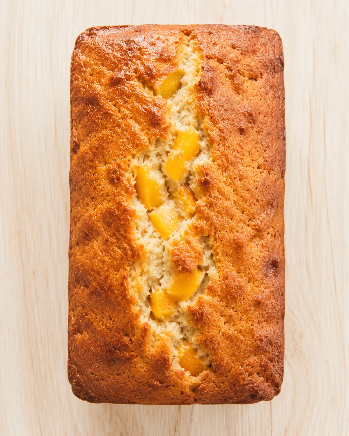 Golden-brown loaf cake with peach chunks visible in a central crack on a light wood surface.